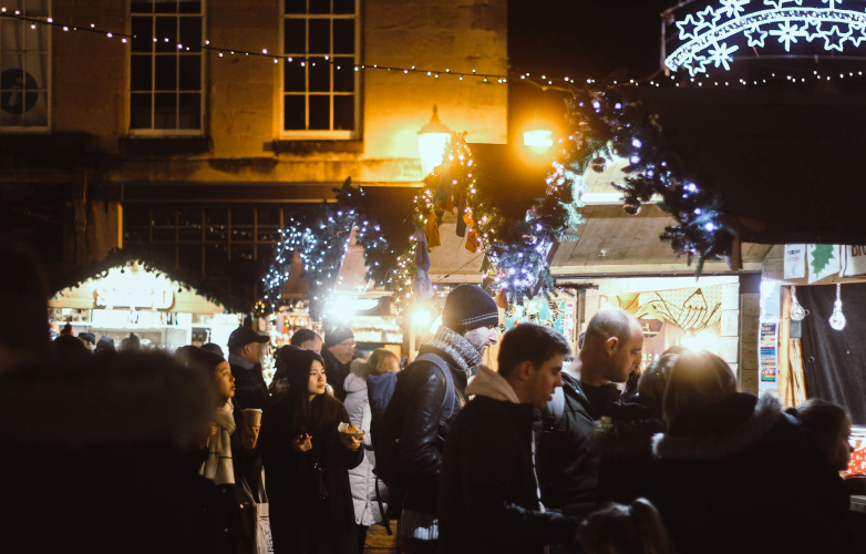 Nighttime photo with christmas lights strung over wooden cabins and people standing to look at the gifts on sale inside