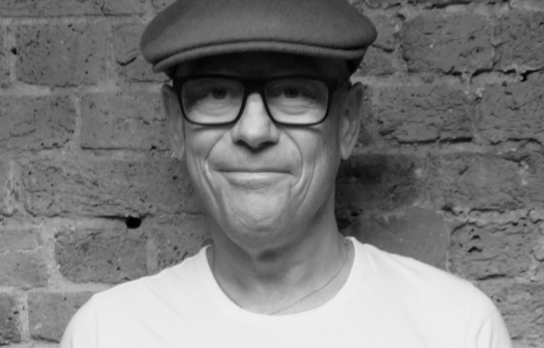Close up of Eric standing against a brick wall looking directly at the camera he wears a flat cap and glasses and is smiling at us