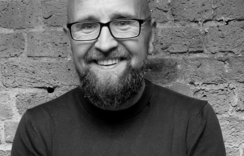 Gordon stands in front of a brick wall he has a broad smile with a thick beard and wears glasses 