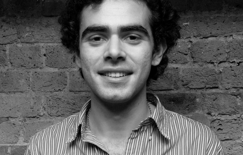 Luis stands in front of a brick wall he has thick short black hair and wears a stripey shirt