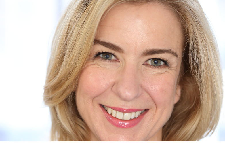 A woman looking directly at the camera smiling she has shoulder length blond hair and wears a black top