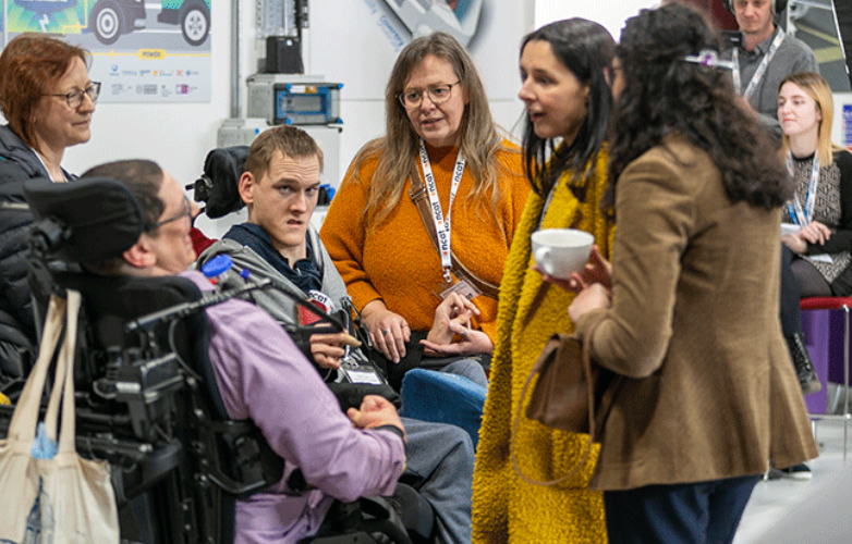 Two male wheelchair users and four women standing in a group at the NCAT launch they wear lanyards round their necks and are chatting and drinking coffee