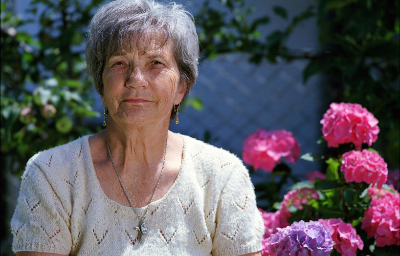 Image shows an older woman with short hair outside by some pink flowers she is smiling at the camera