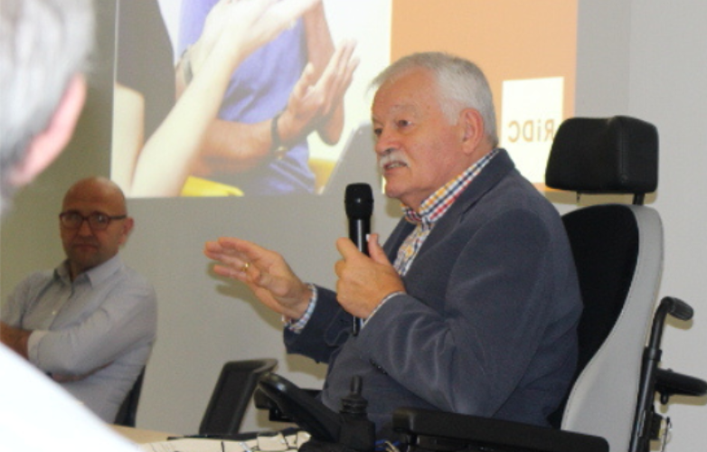 Side on view of Phil sitting in his electric wheelchair holding a microphone addressing an audience Phil has grey short hair and is wearing a dark grey suit jacket with a colourful checked shirt
