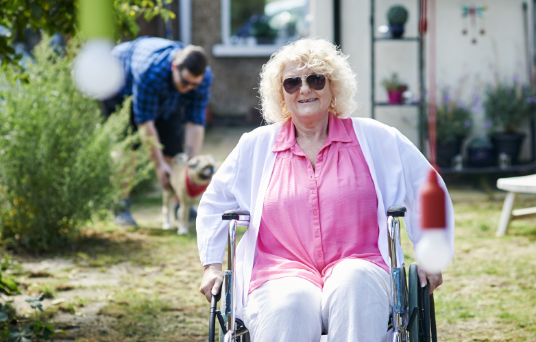 Picture of a woman using a wheelchair in her garden. In the background there is a man playing with a dog. It is a warm and sunny day.