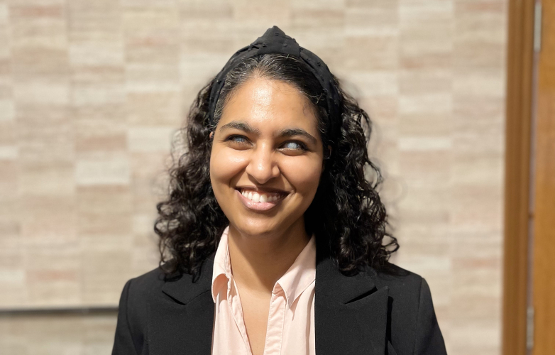 Masuma stands outside smiling at the camera she wears a shirt and suit jacket