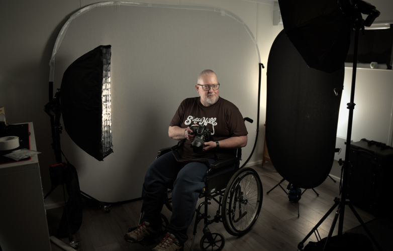Richard sits in his wheelchair in front of the lights on a photographic shoot. He holds his camera and looks towards us