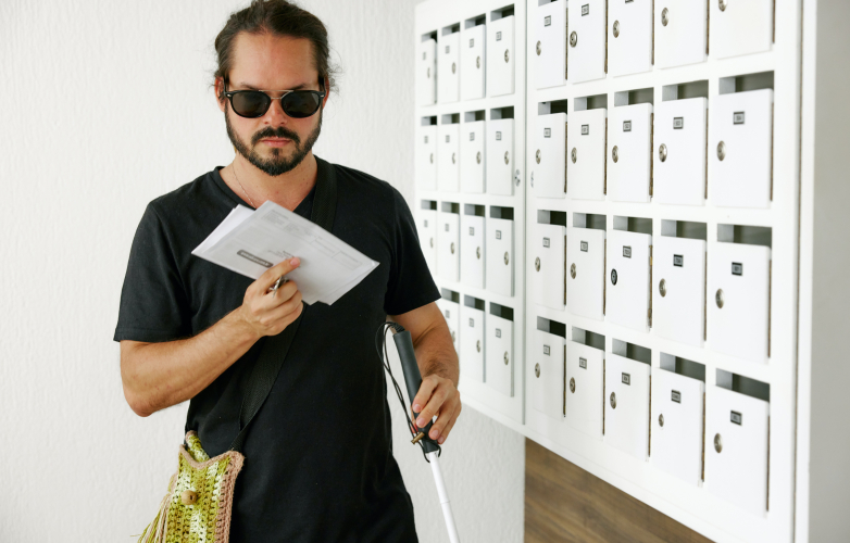 Young man with a visual impairment stands by a bank of letterboxes He is holding two letters in his hand He wears a black tshirt dark glasses and is using a guide cane